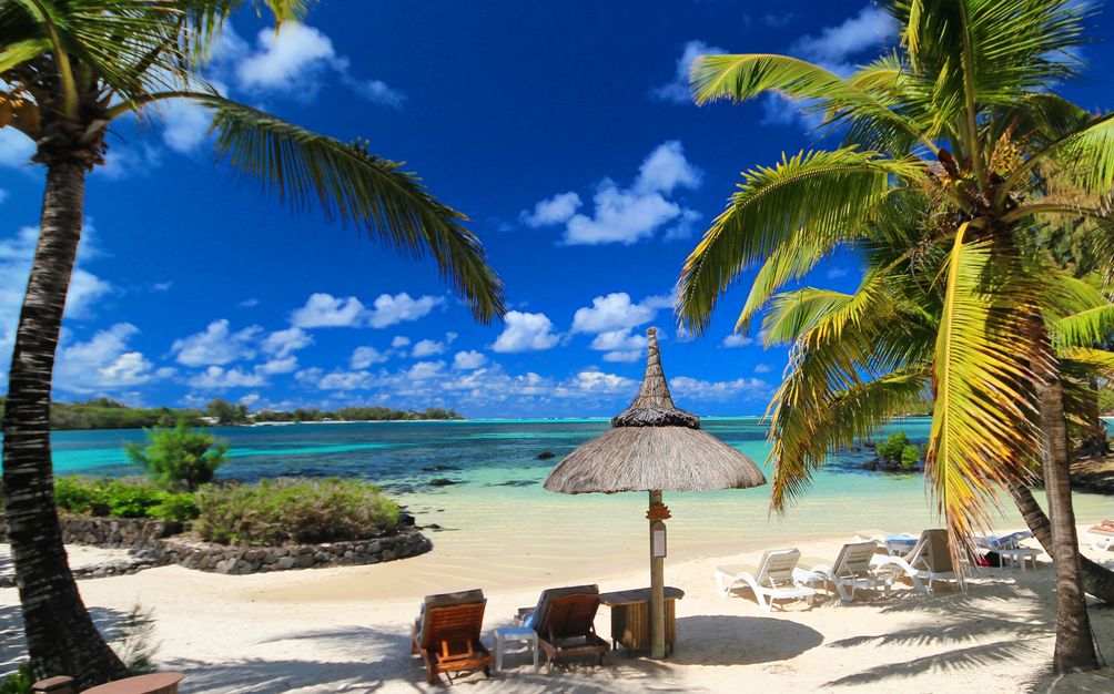 Weißer Sandstrand auf Mauritius mit türkisblauem Wasser, Palmen, einem Sonnenschirm und mehreren Liegen. 